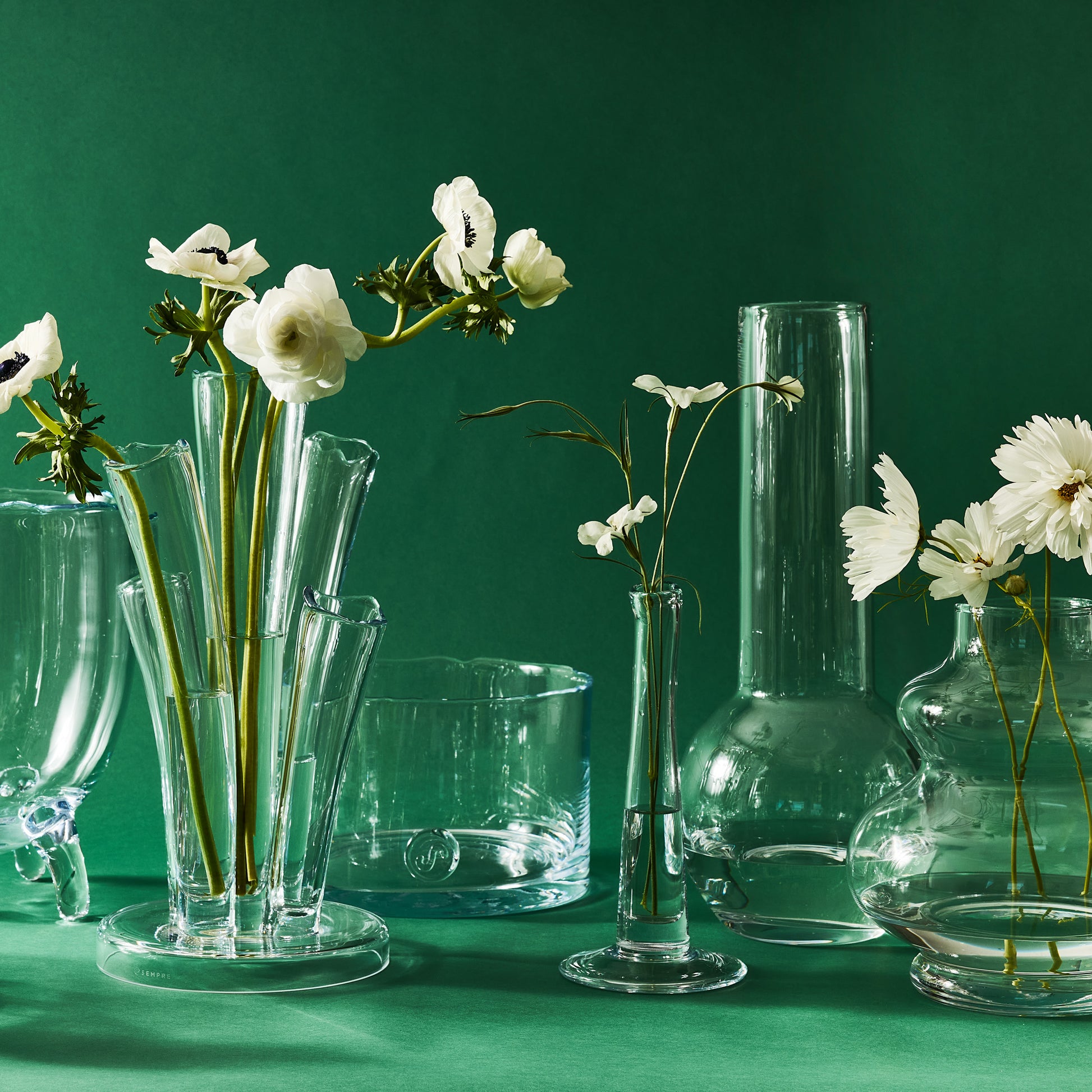 Collection of clear glass vases, including the Sylvia Glass Vase, with white flowers against a green background.