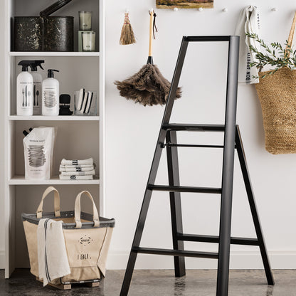 White shelf with HG Tart Kumquat cleaner bottles, a black step ladder, and a woven basket with greenery.