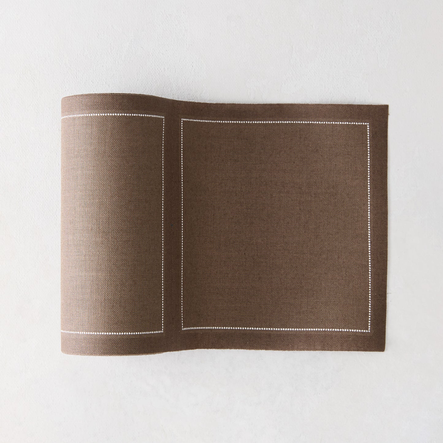 Taupe brown cotton placemat with white stitched border, partially rolled on a white surface.
