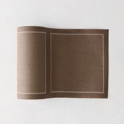 Taupe brown cotton placemat with white stitched border, partially rolled on a white surface.