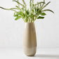 Hand-blown taupe glass vase with a sleek, geometric silhouette and curved edges, 15 inches tall, filled with green stems.