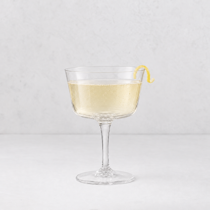 Clear glass coupe cocktail glass filled with sparkling pale yellow liquid and a lemon twist garnish.