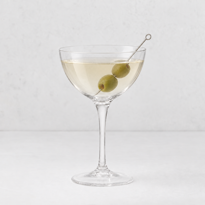 Tavola martini glass with a wide, shallow bowl and two green olives on a metal pick against a white background.