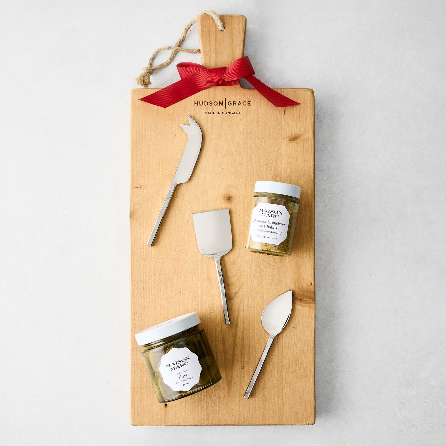 Wooden charcuterie board with red ribbon, silver cheese knives, Maison Marc mustard, and cornichons.