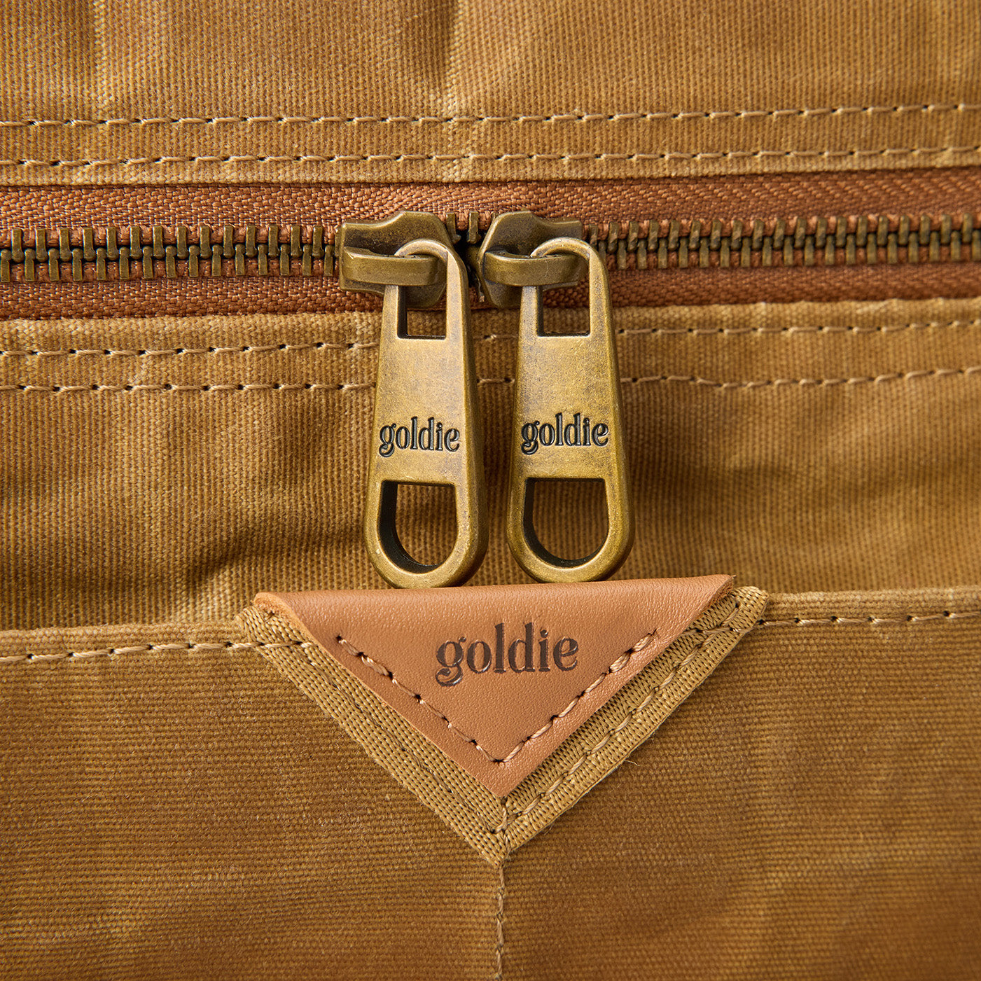 Close-up of Goldie Picnic Bag's waxed canvas, showing two gold "goldie" zipper pulls and a leather "goldie" tag.