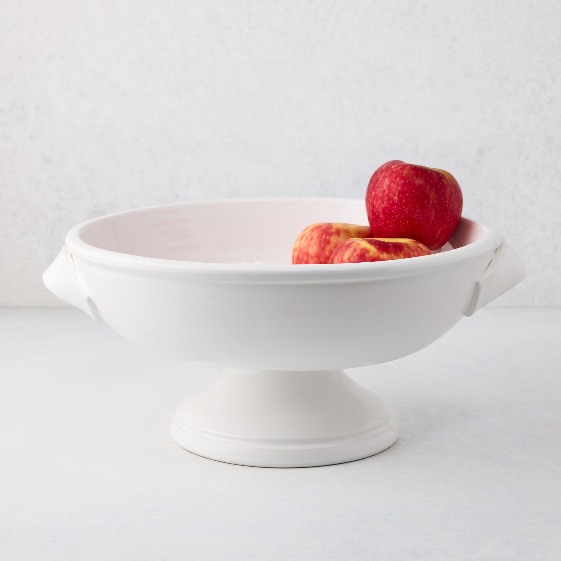White Tivoli ceramic pedestal serve bowl with two handles, holding red apples.