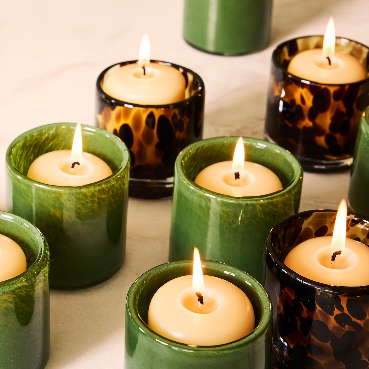 Tortoise and green glass votive candle holders with lit tealights on a light surface.