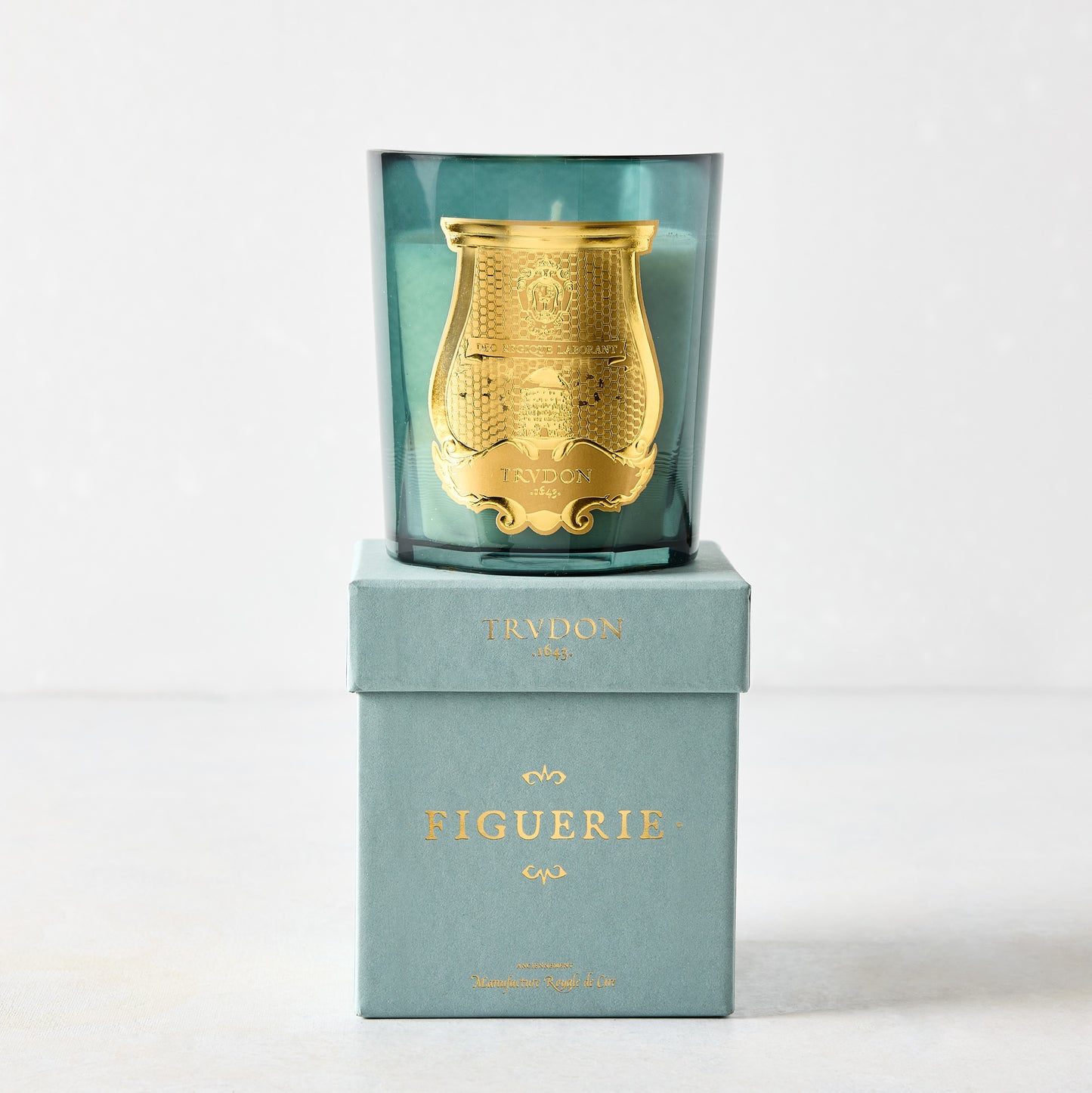 Trudon Figuerie scented candle in a teal glass vessel with a gold seal, on its matching teal box.
