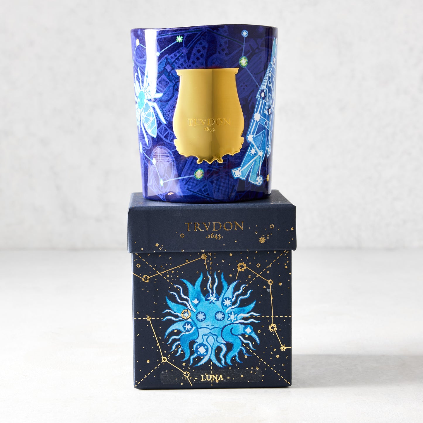 Trudon Luna Holiday Edition scented candle in a blue glass vessel with gold celestial illustrations, atop a matching box.