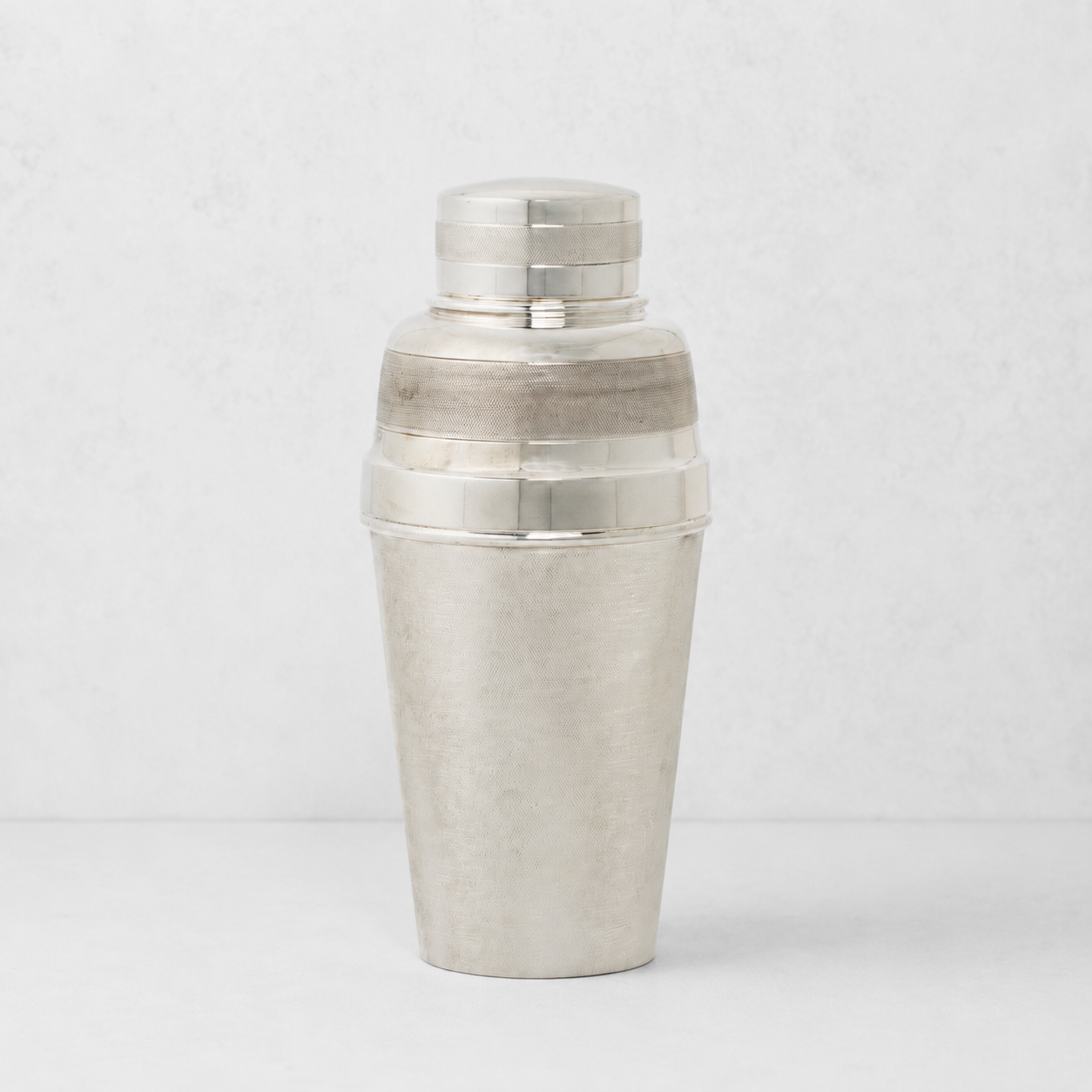 Vintage silverplate cocktail shaker with a textured finish and polished bands against a light gray background.
