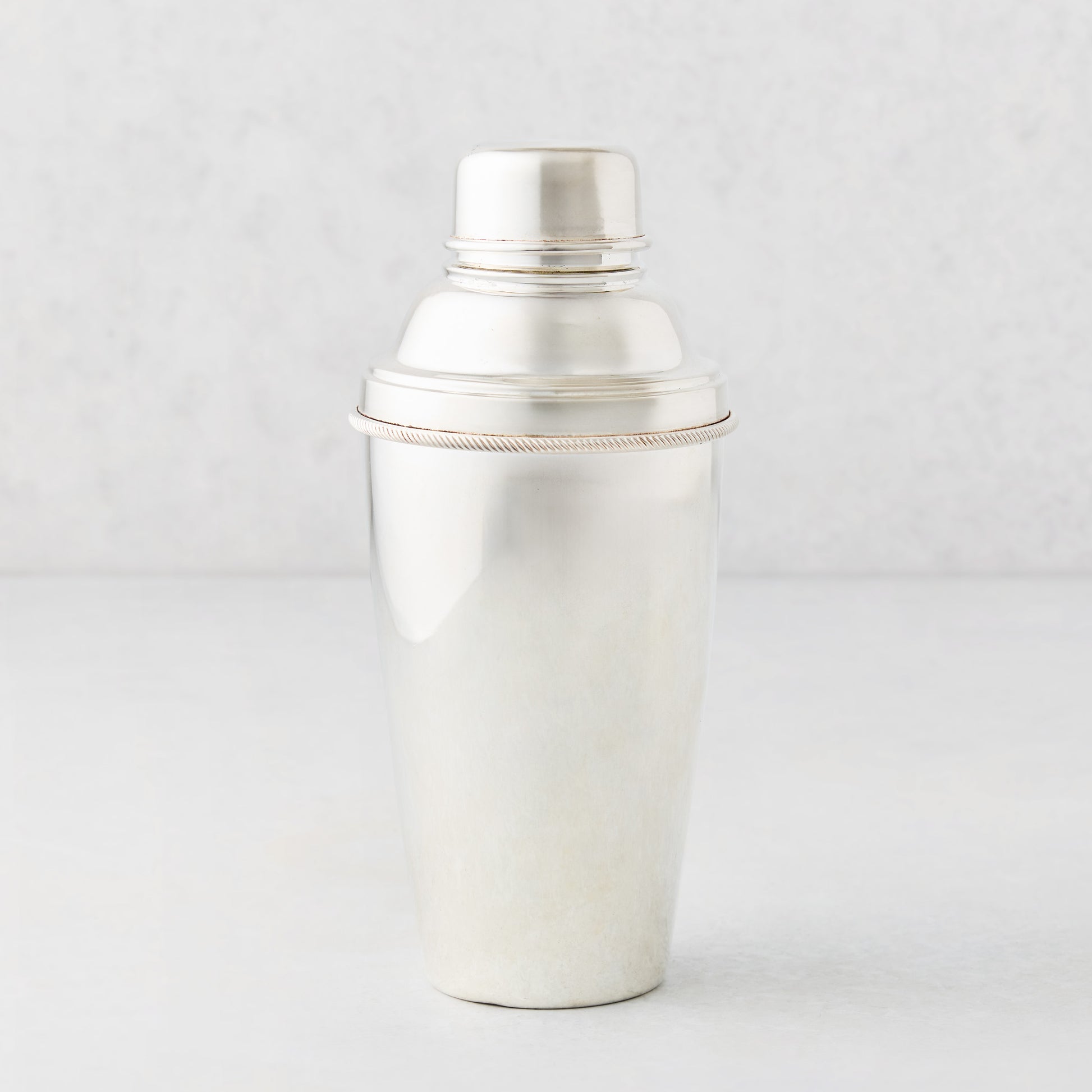 Vintage silverplate cocktail shaker with a smooth finish and rope-detail rim on a light gray background.