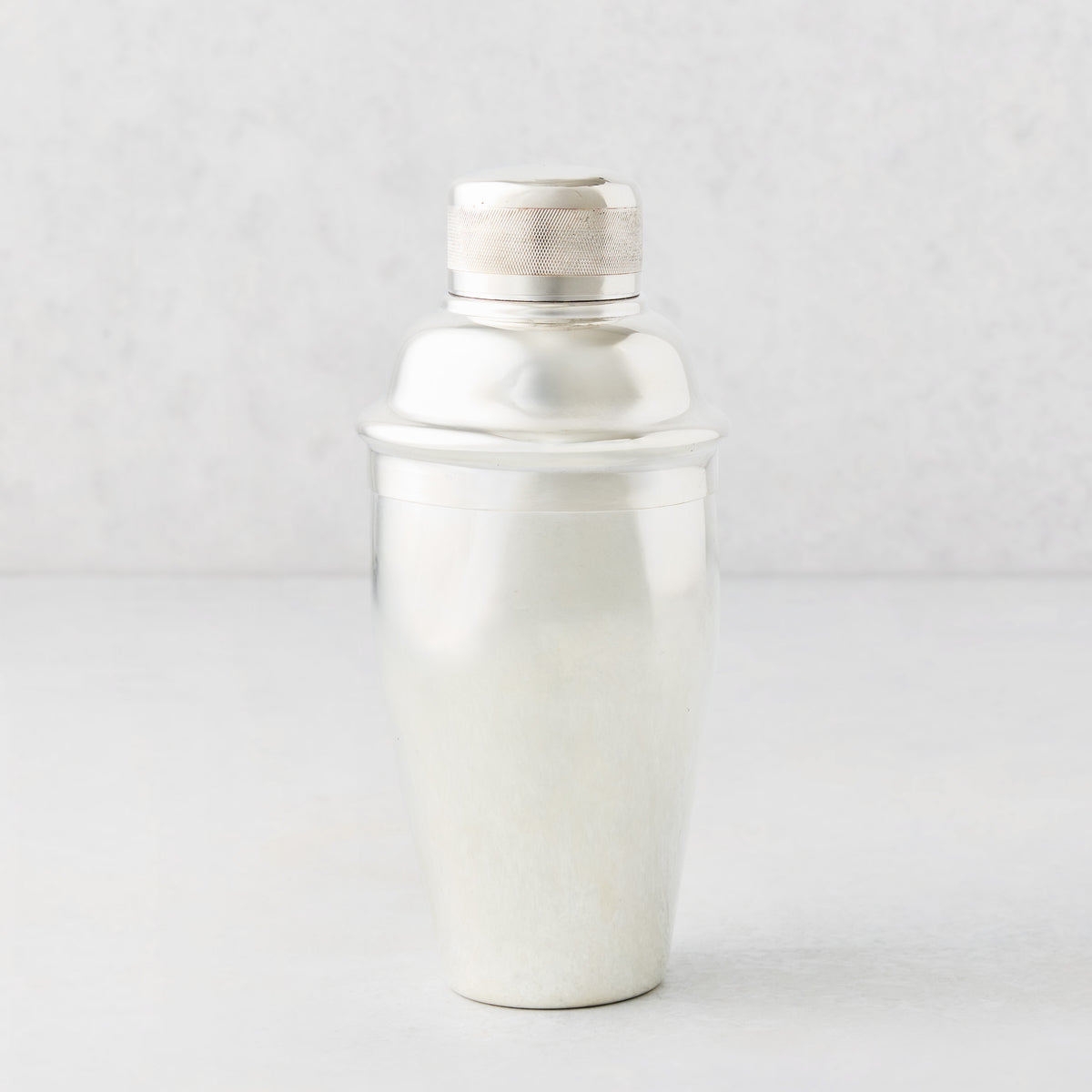 Vintage silverplate cocktail shaker with a smooth body and textured ridged rim cap on a light gray background.