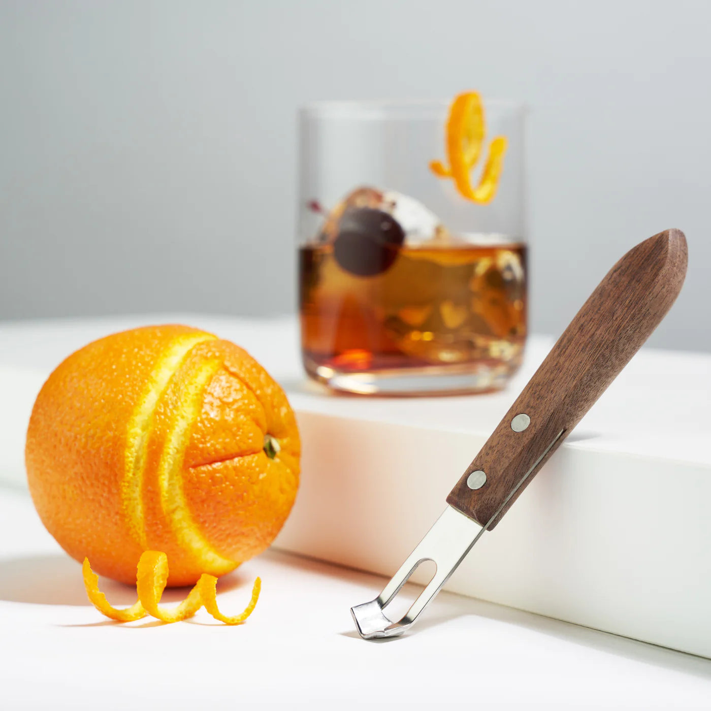 Walnut wood channel knife with stainless steel blade, next to a peeled orange and a cocktail.