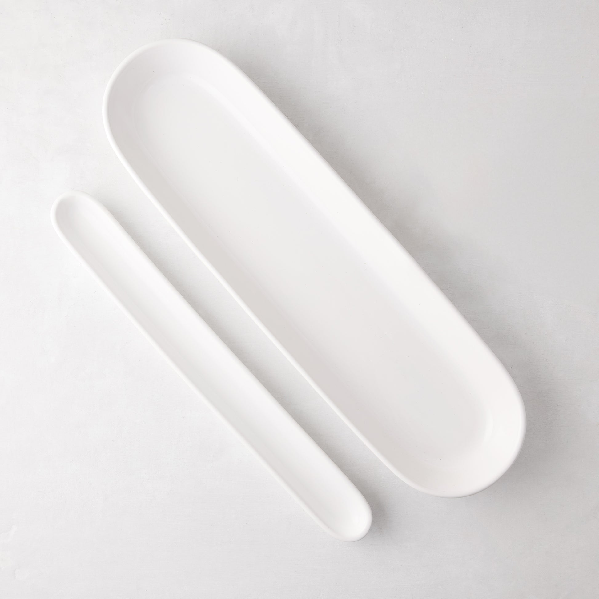 Two white ceramic oval baguette serving trays, one long and one shorter, on a light background.