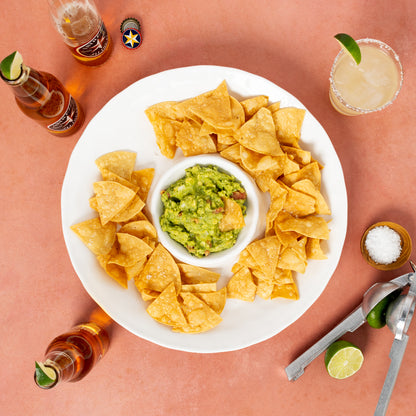 White ceramic 15"Dia chip and dip platter with guacamole and tortilla chips, surrounded by beer, margarita, and limes.