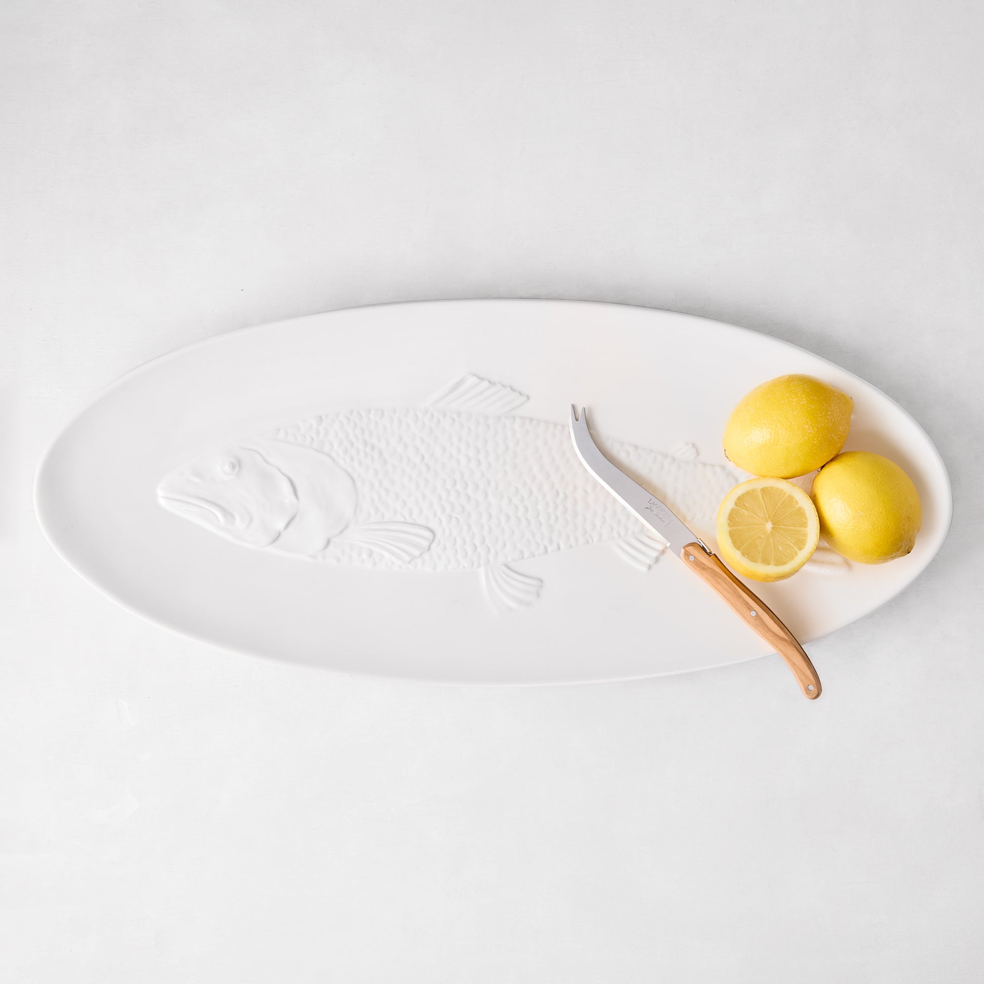 White ceramic oval fish serving platter with embossed fish design, a cheese knife, and lemons.