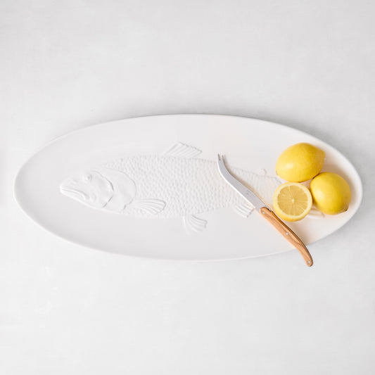White ceramic oval fish serving platter with embossed fish design, a cheese knife, and lemons.