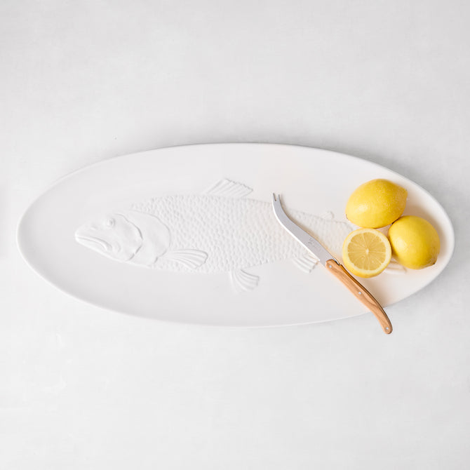 White ceramic oval fish serving platter with embossed fish design, a cheese knife, and lemons.