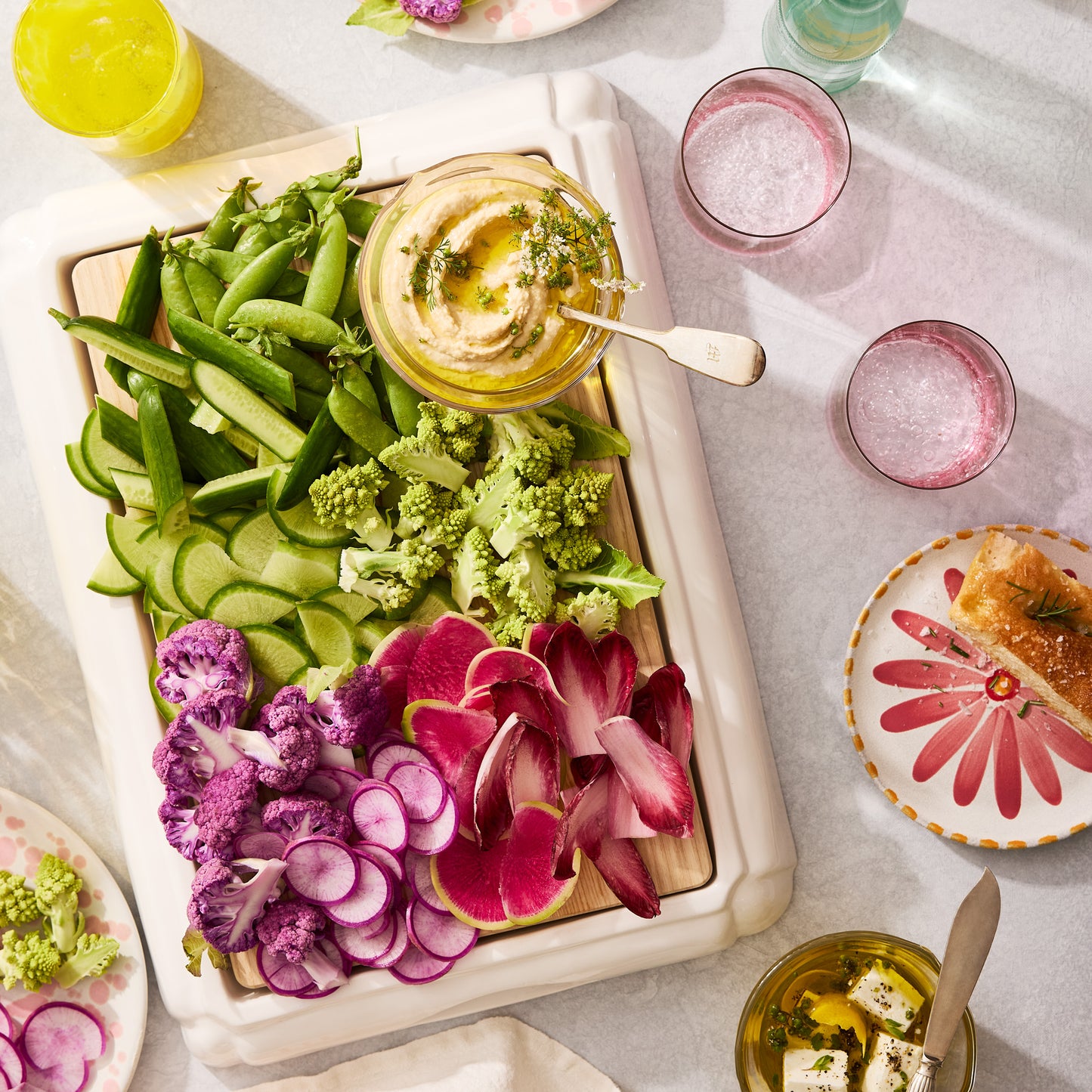 White ceramic handled cutting board with an oak wood surface, serving colorful vegetables and hummus.