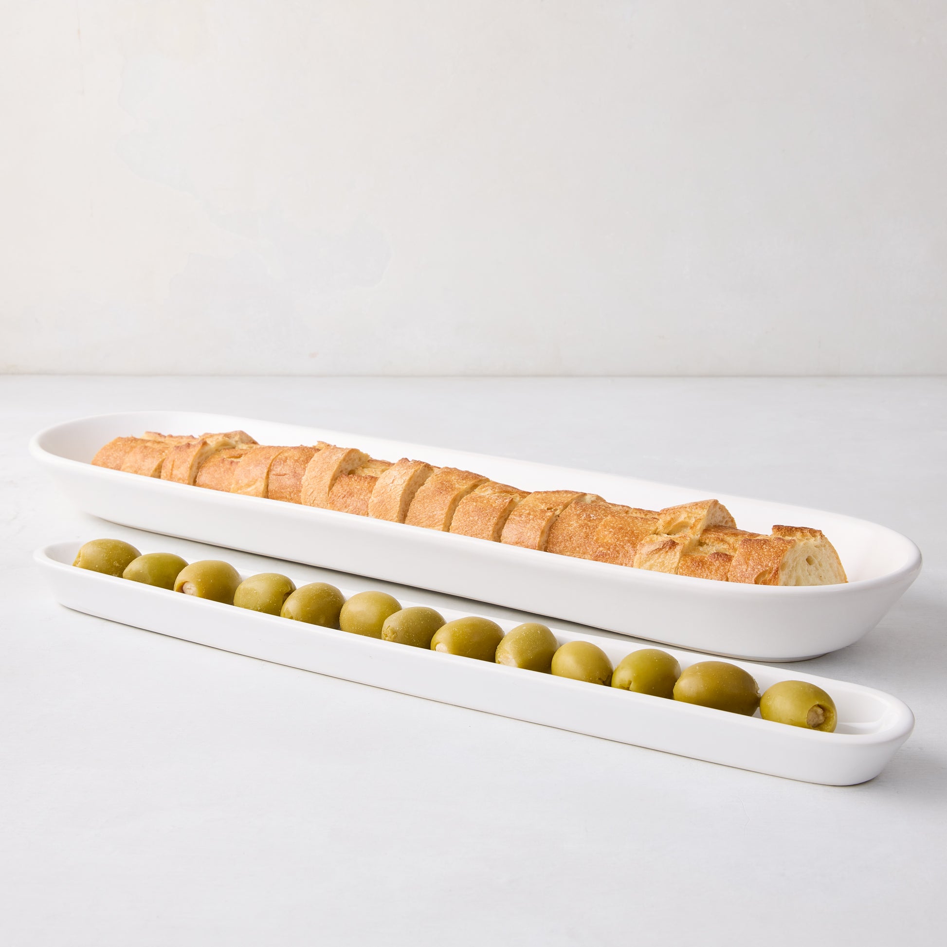 White ceramic olive serving tray, 16"L, with green olives in front of a larger baguette tray.