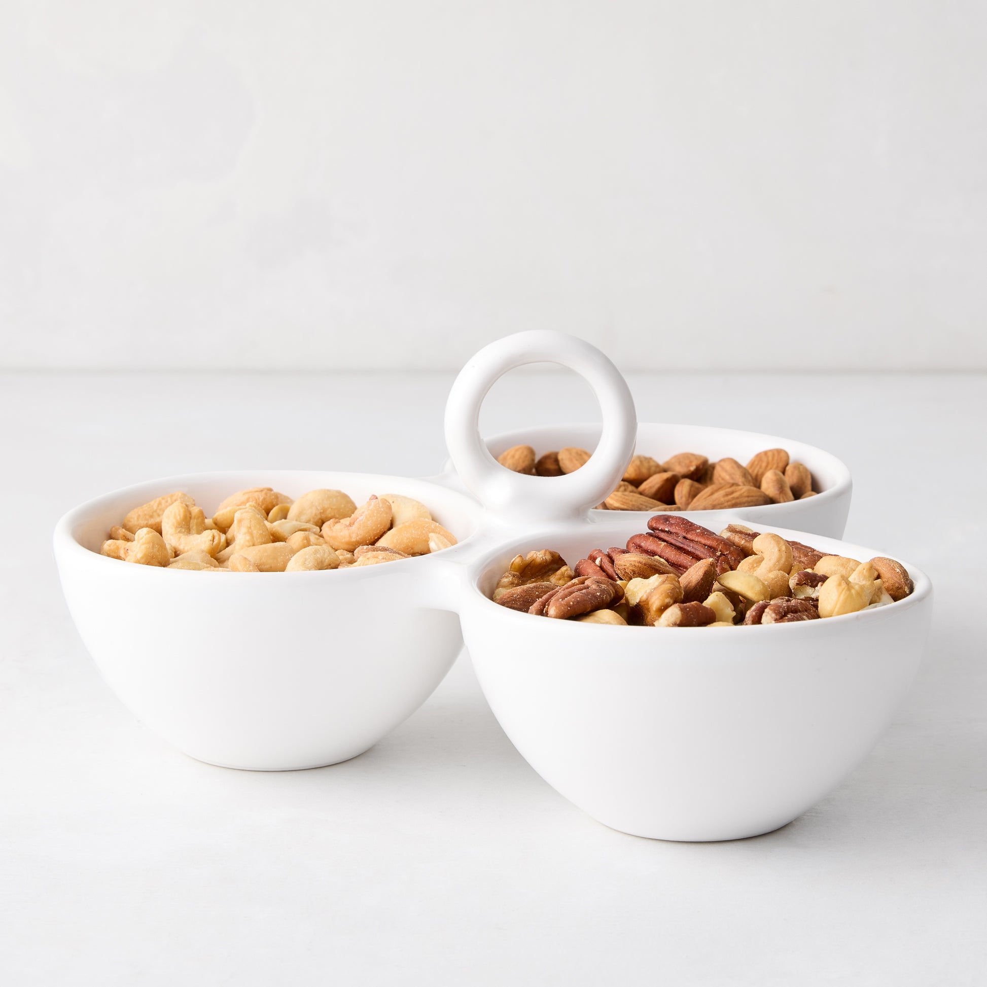 White ceramic triple serve bowl with handle, filled with cashews, almonds, and pecans.