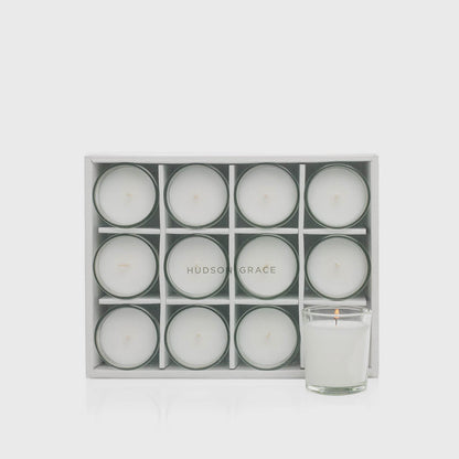 Set of 12 white paraffin votive candles in clear glass cups, one lit, in a white storage box.