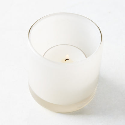 White glass votive candle holder with a lit tealight candle inside.