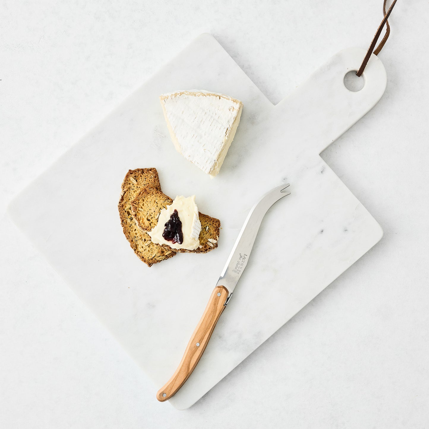 White marble square serve board with handle and leather strap, holding cheese, cracker, and knife.
