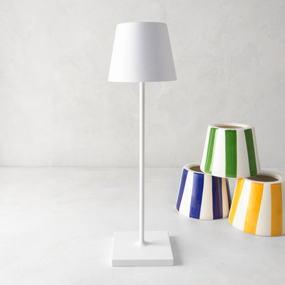 White Poldina rechargeable lamp with a sleek, modern design, standing next to colorful striped planters.