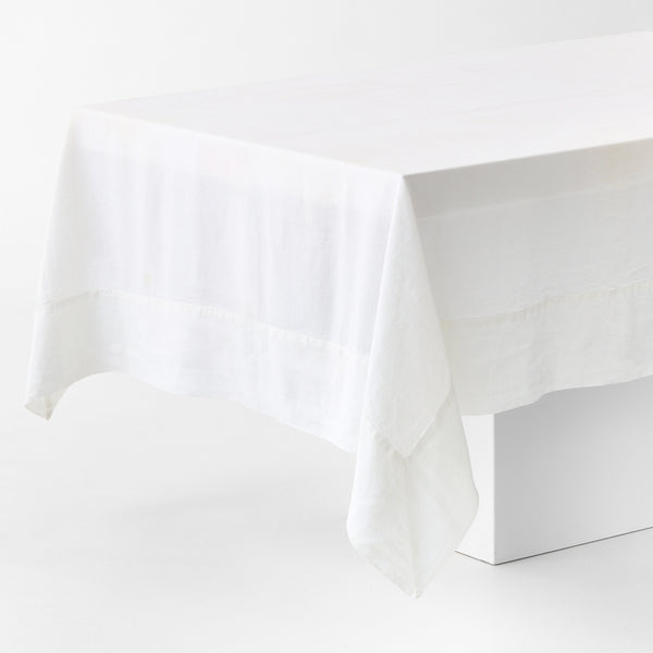 White hand-dyed 100% linen tablecloth with a wide border and mitered corner detailing draped over a table.