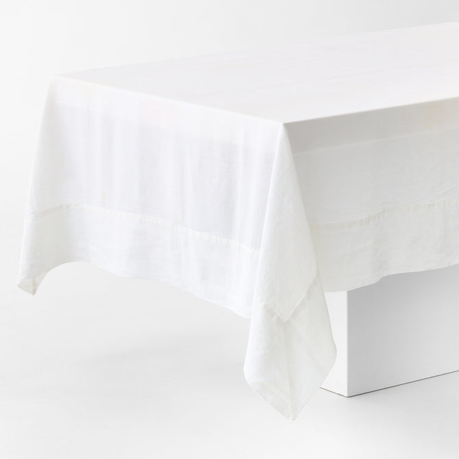 White hand-dyed 100% linen tablecloth with a wide border and mitered corner detailing draped over a table.