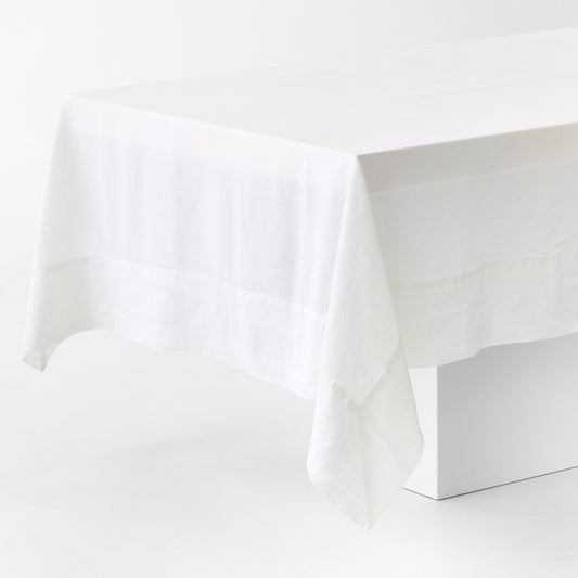 White hand-dyed 100% linen tablecloth with a wide border and mitered corner detailing draped over a table.