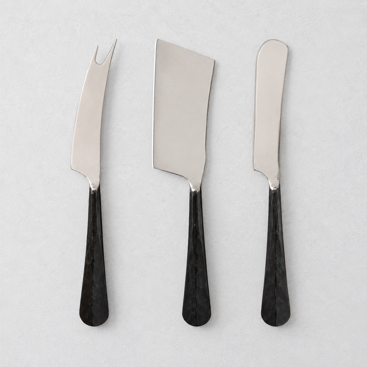 Three-piece cheese knife set with stainless steel blades and black, hand-forged, hammered metal handles.