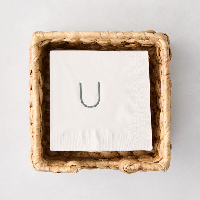 Square woven water hyacinth cocktail napkin holder with white monogrammed napkin inside.