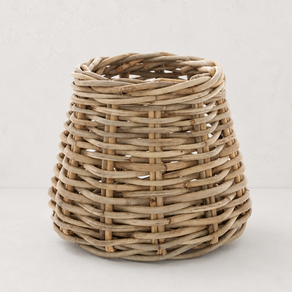 A natural woven wicker basket with a tapered, rounded shape, shown on a clean, light off-white background.