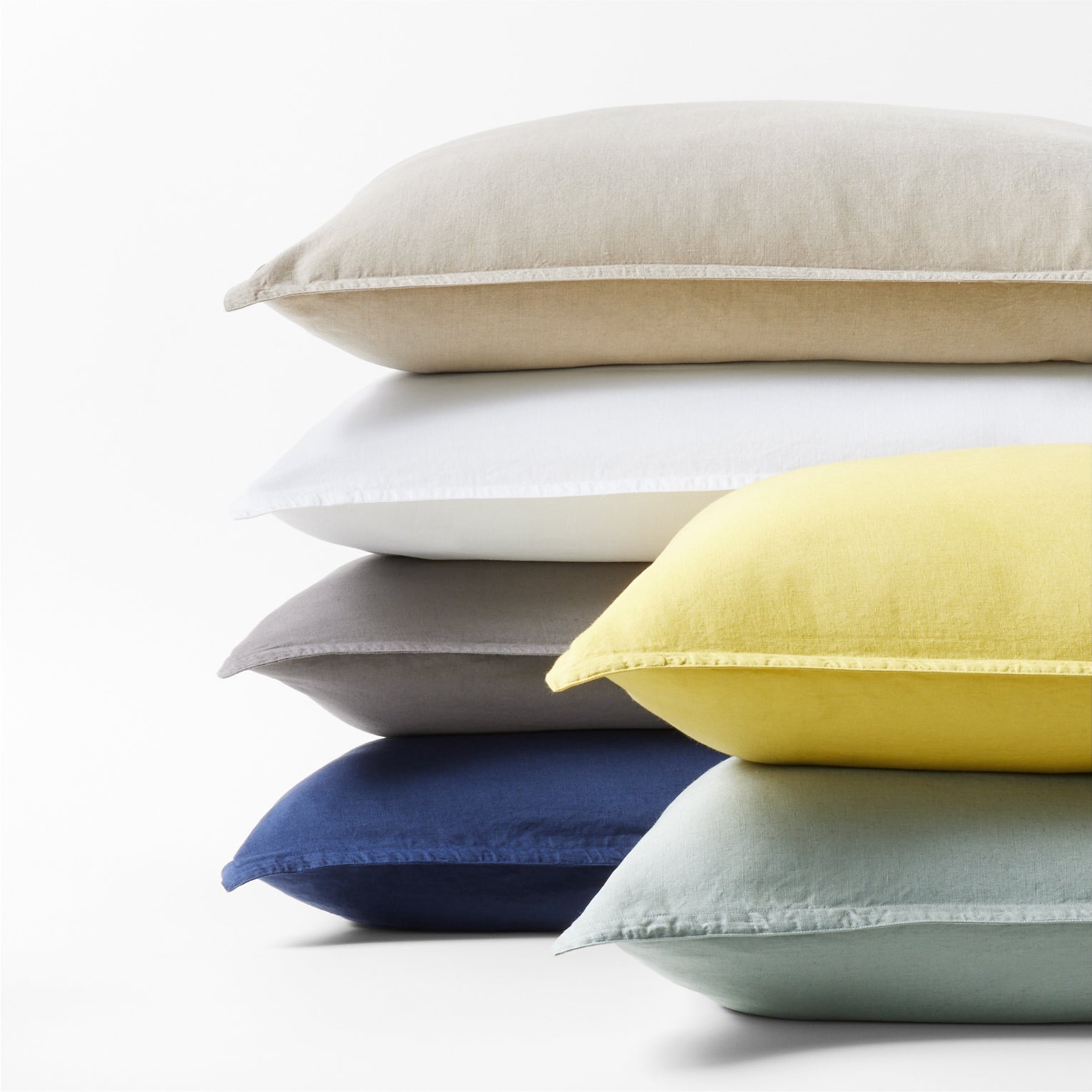 Stack of washed linen pillow shams in yellow, beige, white, grey, navy, and light green.