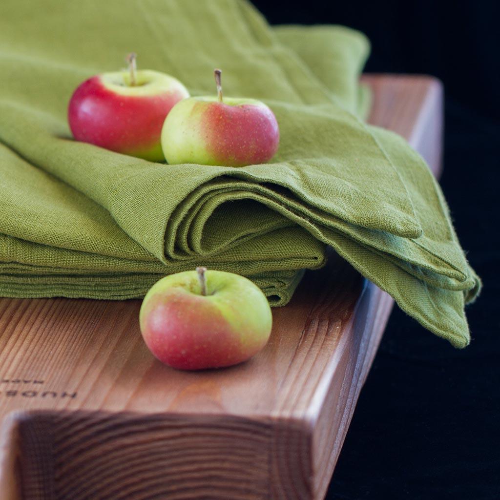 Aloe Green HG Signature Hand-dyed Linen Napkin, folded on a wooden board with three small apples.