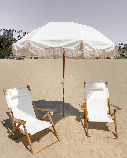 Two Antique White Reclining Tommy Outdoor Beach Chairs with a fringed white umbrella on sand.