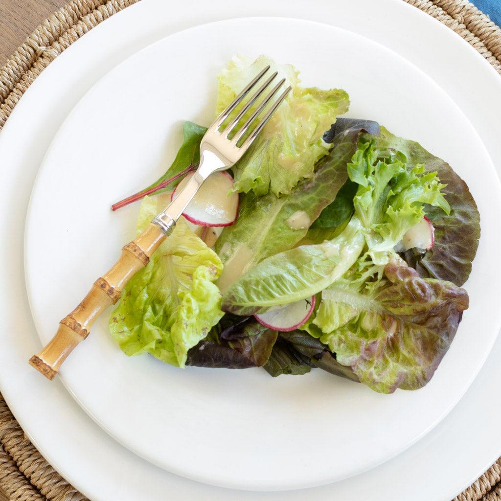 Bamboo 5-piece flatware set with a fork featuring a natural bamboo handle and 18/10 stainless steel tines, resting on a white plate with salad.