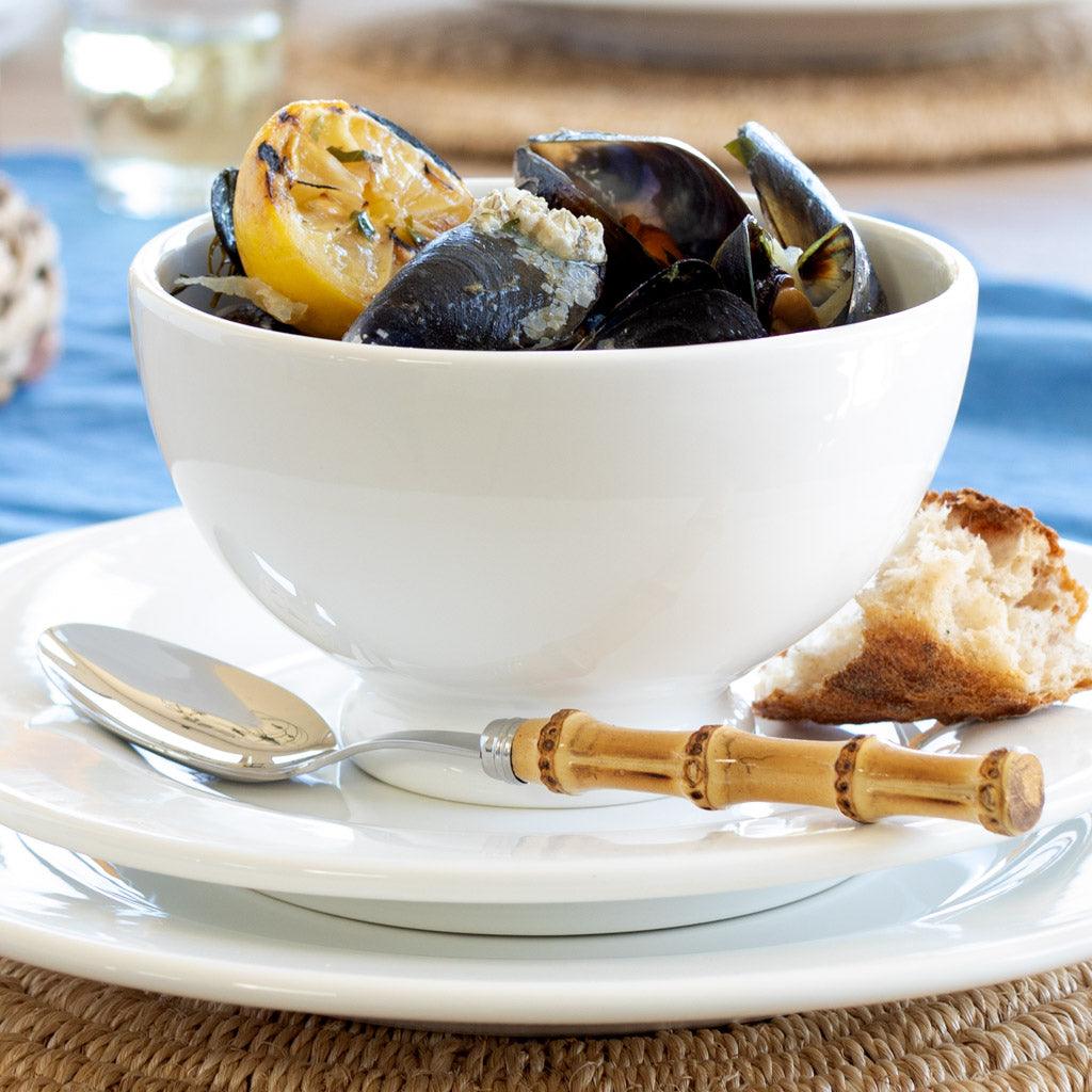 White bowl of mussels with lemon, a slice of bread, and a silver spoon with a bamboo handle.