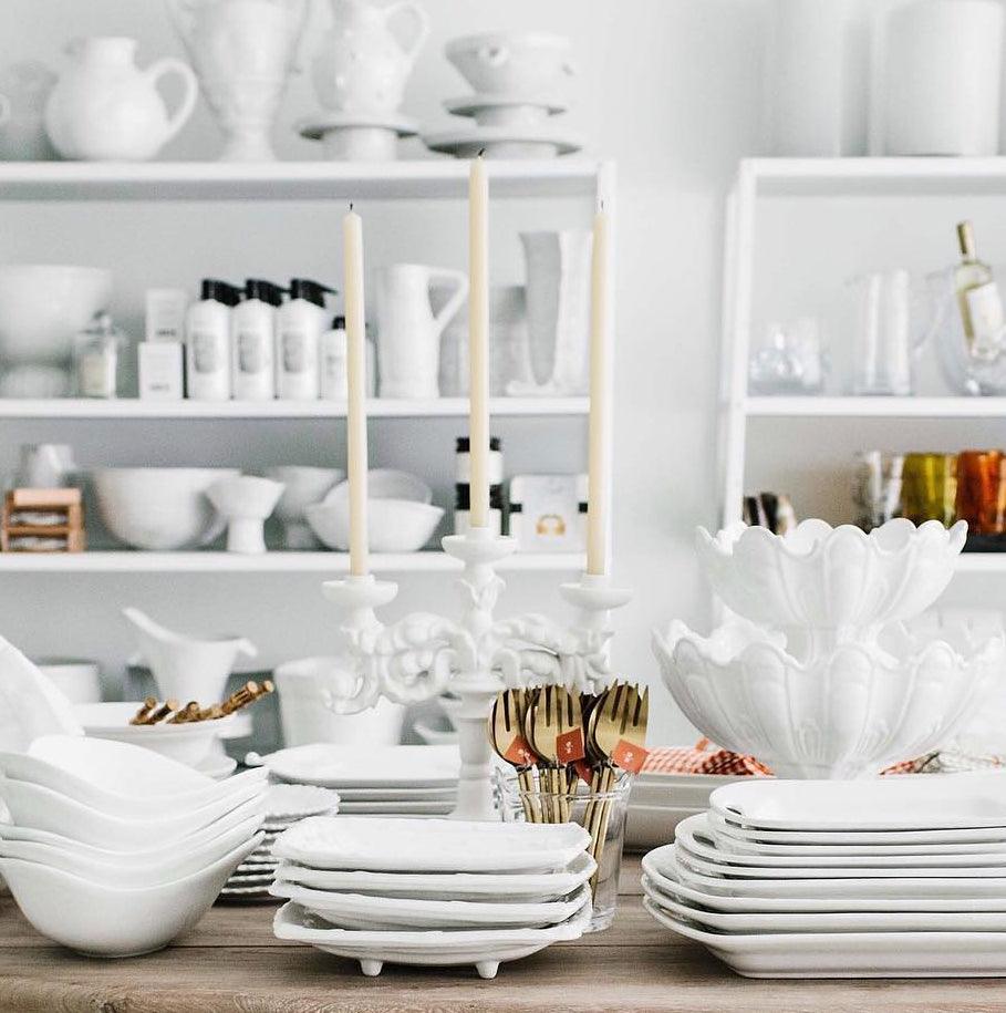 Baroque matte white ceramic candelabra with three lit candles, surrounded by white dishware on a wooden table.