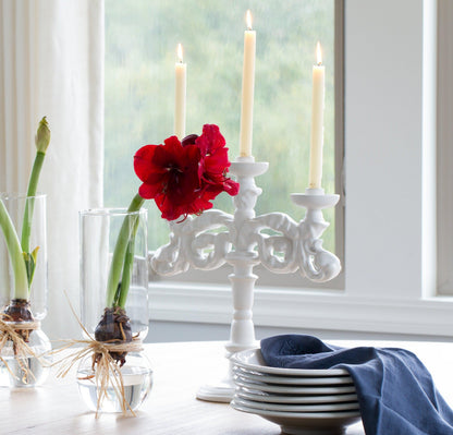 Baroque matte white ceramic candelabra with three lit candles, next to red amaryllis and stacked plates.