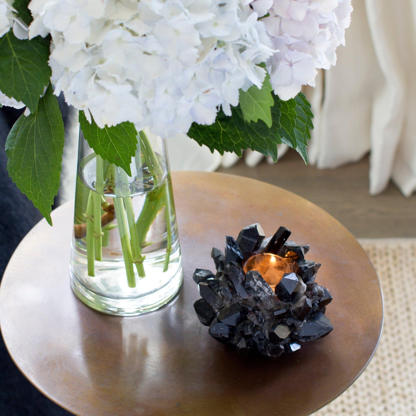 Black classic quartz votive candle holder with a lit candle, next to a vase of white hydrangeas.