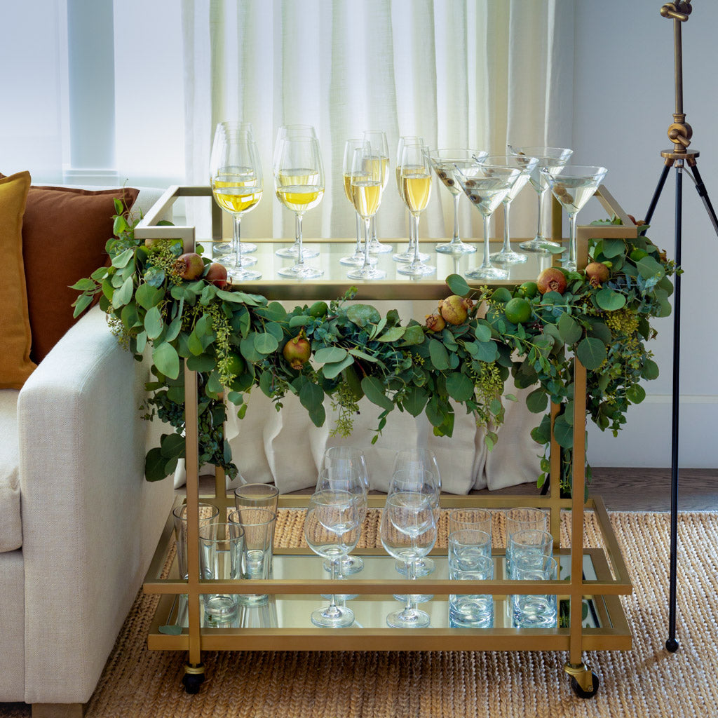 Antique brass bar cart with two mirrored shelves, decorated with a green garland and various glassware.