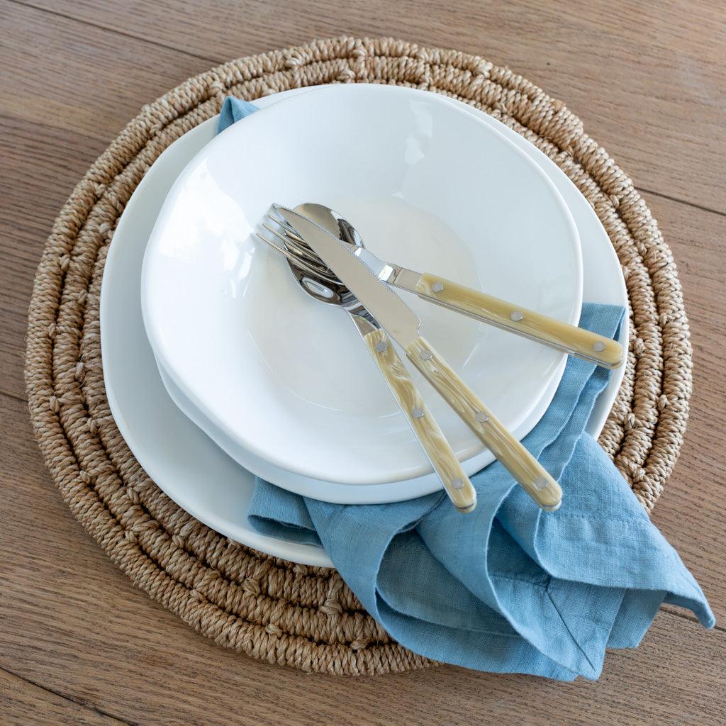 Cloudy HG Signature hand-dyed linen napkin, muted blue-gray, folded under white plates on a woven placemat.