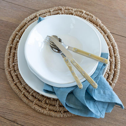 Cloudy HG Signature hand-dyed linen napkin, muted blue-gray, folded under white plates on a woven placemat.