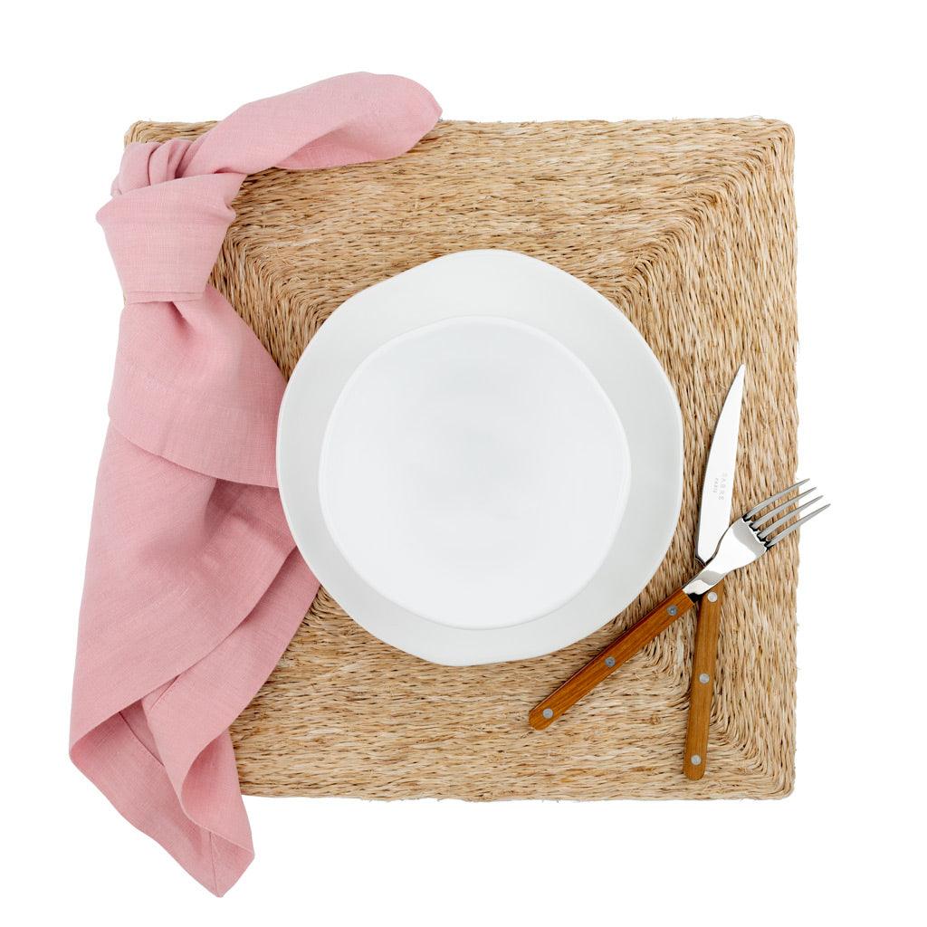 Clover Pink HG Signature hand-dyed linen napkin, knotted on a woven placemat with a white plate and cutlery.