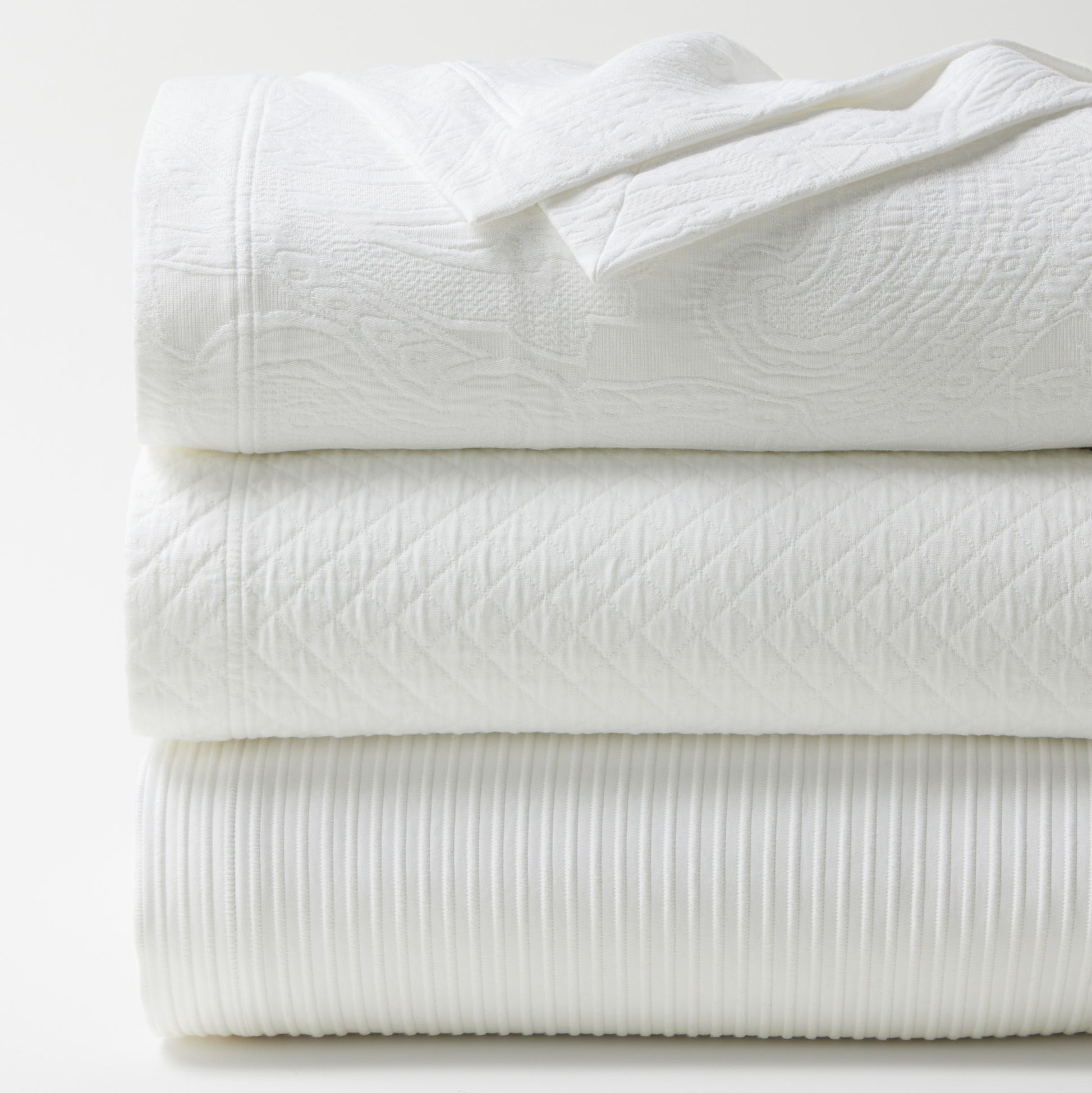 Stacked white cotton matelassé coverlets with diamond, paisley, and ribbed textures.