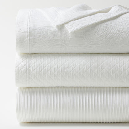 Stacked white cotton matelassé coverlets with diamond, paisley, and ribbed textures.