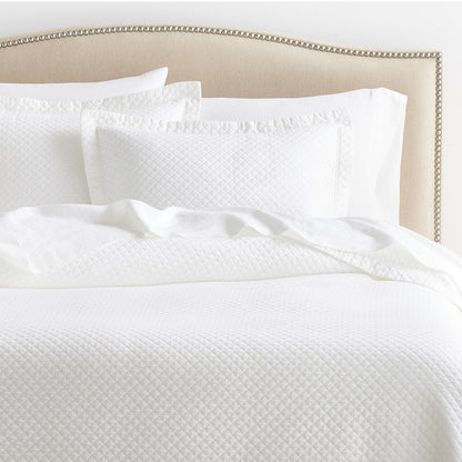 Diamond White Cotton Matelassé Pillow Shams and quilt on a bed with a beige upholstered headboard.